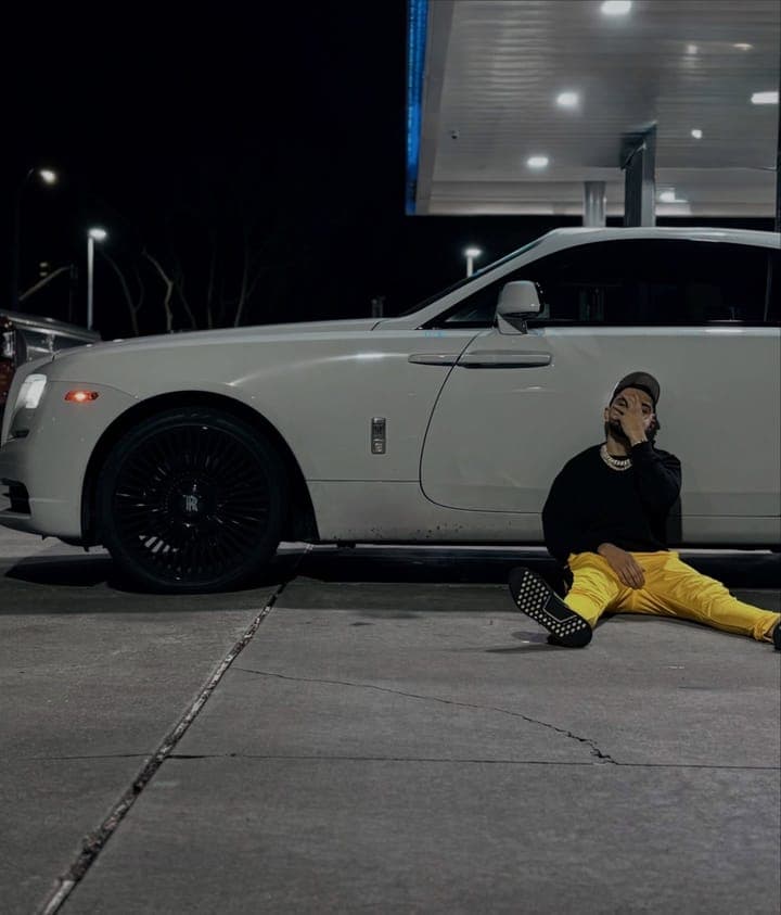 Sitting on the ground against a white Rolls-Royce under a gas station canopy at night, in a black sweatshirt, yellow trousers and a Cuban link chain, one hand over his face