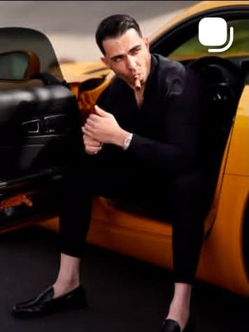 A man in an all black suit seated in the open door of an orange sports car