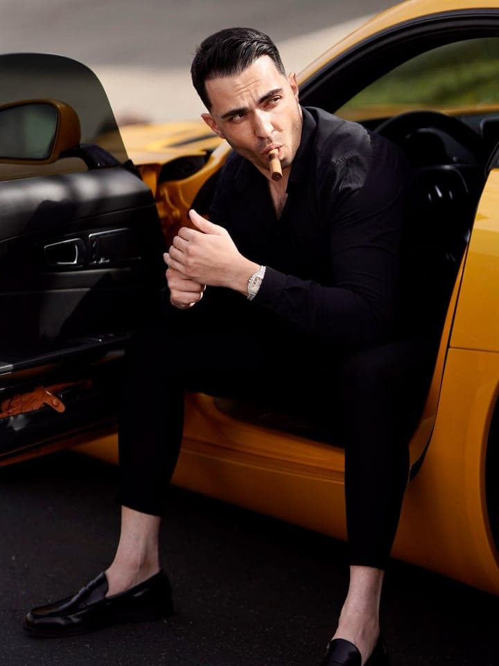 A man in an all black suit seated in the open door of an orange sports car, a cigar in his mouth