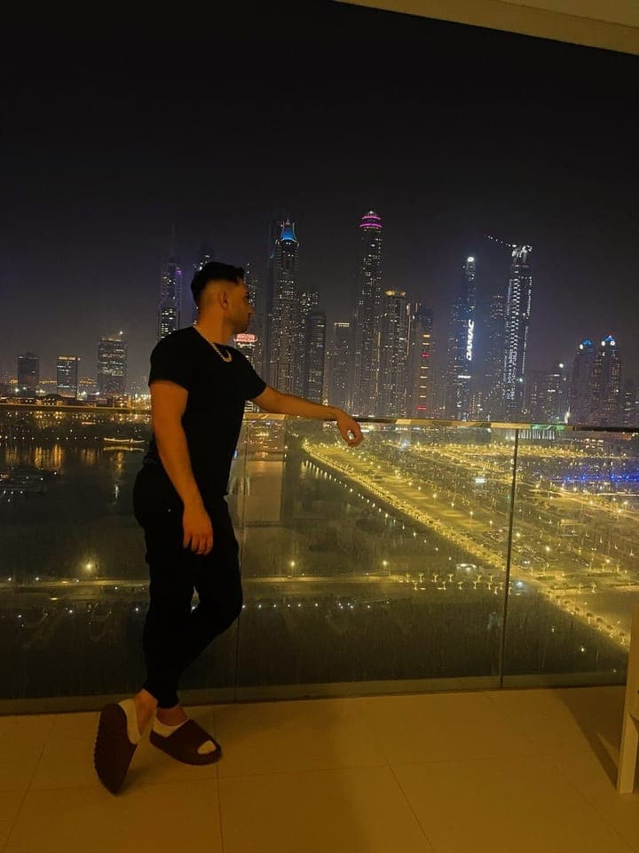 Leaning on a balcony rail at night above the lit boulevards of Dubai Marina, in all black with a Cuban link chain